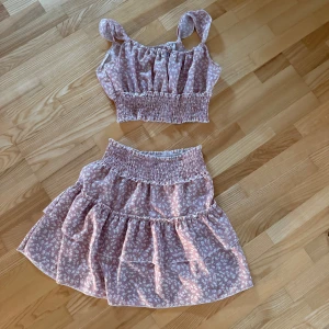 volangkjol med topp - Supersöt rosa kortkjol med små vita blommor och volanger. Kjolen har smock i midjan för en bekväm passform och är perfekt för varma dagar. Tunt och luftigt material som känns lätt på kroppen.