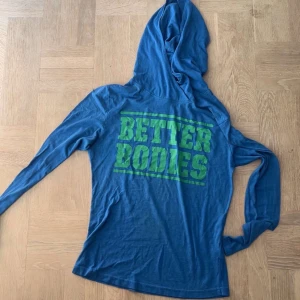 Blå hoodie från Better Bodies - Snygg blå hoodie från Better Bodies med stort grönt tryck på ryggen. Långärmad modell med huva, perfekt för träning eller chill. Mjukt material som känns skönt mot huden och ger en avslappnad vibe.