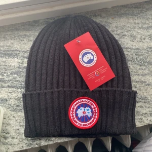 Ribbstickad mössa Canada Goose - Svart ribbstickad mössa från Canada Goose med klassisk rund logotyp-patch framtill. Tjockt uppvikt kant och stilren design som håller dig varm. Perfekt för kalla dagar och snygg till alla outfits. Liten i storleken men går att töja ut
