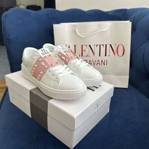 Valentino Rockstud vita sneakers - Snygga vita Valentino Rockstud sneakers med ljusrosa detaljer och ikoniska nitar. Skorna har rund tå, platt sula och är tillverkade i skinn. Perfekta för dig som vill ha en lyxig och trendig look med en touch av rosa och coola metalldetaljer.