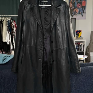 Svart lång skinnrock Giorgio Valenti - Säljer en svart lång skinnrock från Giorgio Valenti i äkta lambskin läder. Jackan har klassisk krage, knäppning med fyra knappar framtill och raka långa ärmar. Snygg minimalistisk look som ger en cool vibe till vilken outfit som helst.