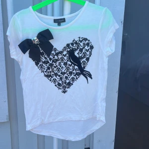 Vit t-shirt med hjärta och rosett KappAhl - Vit t-shirt från KappAhl med korta ärmar, dekorerad med ett stort svart hjärta i blommigt mönster och en svart fågel framtill. Snygg svart rosett med paljettdetalj på axeln. Mjuk och lätt bomullskänsla, perfekt till sommaren.