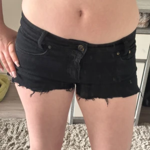 Svarta jeansshorts från Sweet Poison - Supersnygga svarta jeansshorts från Bik Bok tough love med låg midja och råa, fransiga kanter. Klassisk femficksmodell med guldiga nitar och knapp. Perfekta för sommarens alla äventyr. Sitter tight och har en edgy vibe. Står storlek M men känns mer som XS eller S
