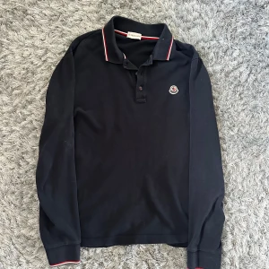Svart långärmad pikétröja Moncler - Svart långärmad pikétröja från Moncler med klassisk krage och knäppning framtill. Tröjan har röda och vita ränder på krage och ärmslut samt Moncler-logga på bröstet. Tillverkad i mjuk bomull, perfekt för en clean och sportig look.