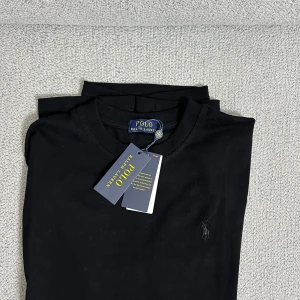 Svart t-shirt Polo Ralph Lauren - Svart t-shirt från Polo Ralph Lauren med klassisk passform och rund hals. Diskret broderad logga på bröstet och mjukt bomullsmaterial. Perfekt basic-plagg med stilren vibe och snygg finish.