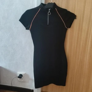 Svart kortklänning med dragkedja - Svart tight kortklänning med korta ärmar och hög krage. Snygg detalj med silverfärgad dragkedja framtill och kontrasterande sömmar längs axlarna. Perfekt för en trendig och sportig look.