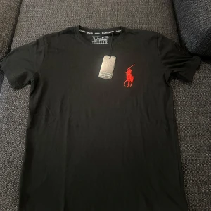 Svart t-shirt från Ralph Lauren - Svart t-shirt från Ralph Lauren med stor röd broderad polospelare på bröstet och svart broderad siffra 3 på ena ärmen. Klassisk passform och rund hals. Tillverkad i mjuk bomull, perfekt för en clean och stilren look.