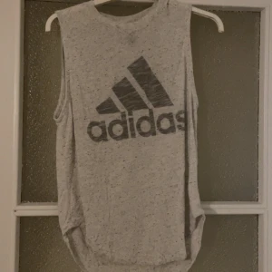 Grå ärmlös linnetopp från Adidas - Snygg gråmelerad linnetopp från Adidas med stort svart logotryck framtill. Modellen är ärmlös och har en avslappnad passform, perfekt för träning eller chill dagar. Tunt och mjukt material som andas.
