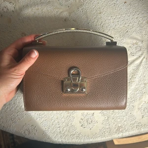 Brun handväska i skinn med metallhandtag - Präglat skinn, klassisk väska från Aigner. Väskan har två fack och ett innerfack med dragkedja, använd fåtal gånger dvs inga tecken på användning.
