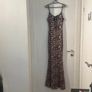 Lång leopardklänning med snörning. Helt ny - Snygg långklänning med leopardmönster i beige och brunt. Klänningen har smala axelband, v-ringning och en cool snörningsdetalj framtill. Materialet känns slätt och följsamt, perfekt för dig som vill sticka ut.