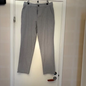 Grå kostymbyxor Jack & Jones Ollie - Snygga grå kostymbyxor från Jack & Jones, modell Ollie Regular. Byxorna har normal passform, bälteshällor, snedställda fickor fram och bakficka med knapp. Perfekta för en clean och stilren look.