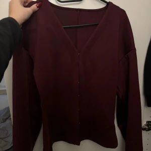 Helt ny vinröd långärmad blus - Säljer en elegant vinröd blus med långa ärmar och v-ringning. Perfekt för en stilren look. Helt ny och aldrig använd. Blusen har en enkel design utan mönster och är gjord i ett mjukt material.