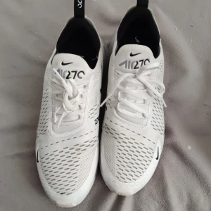 Vita Nike Air Max 270 sneakers - Snygga vita Nike Air Max 270 sneakers med svart logga och synlig luftenhet i hälen. Skorna har en modern design med ventilerande meshmaterial och snörning för bra passform.