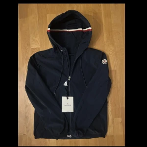 Mörkblå vindjacka från Moncler - Snygg mörkblå vindjacka från Moncler med dragkedja och huva. Jackan har en diskret logga på ärmen och snörning vid huvan. Perfekt för blåsiga dagar och ger en stilren look. Några frågor? Tveka inte på att höra av er.