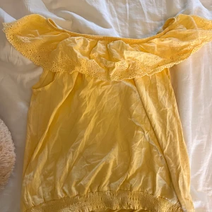 Gul offshoulder topp med spetsdetaljer - Säljer en söt gul offshoulder topp med spetsdetaljer runt halsen. Toppen har en elastisk nederkant och är perfekt för sommardagar. Den är ärmlös och har en luftig passform.