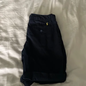 Svarta byxor från Polo Ralph Lauren - Snygga svarta shorts från Polo Ralph Lauren med en klassisk design. Byxorna har en knapp och dragkedja framtill samt en liten broderad logga i gult. Perfekta för en stilren look.