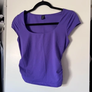 Lila topp - Säljer en snygg lila topp med korta ärmar och rund halsringning. Perfekt för en färgglad look! Toppen har rynkade detaljer på sidorna och dubbelt tyg vid brösten. De noppror som syns på bilderna är borta då jag använt en avnoppringmaskin 🤗 Toppen är använd 2-3 gånger. Önskar du veta exakta mått? Tveka inte att höra av dig!