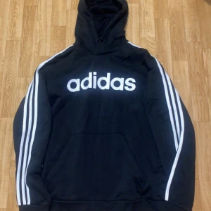 Adidas hoodie (tröja) - Säljer en klassisk svart hoodie från Adidas med den ikoniska loggan i vitt på bröstet. Tröjan har de välkända tre ränderna längs ärmarna och en praktisk magficka. Perfekt för en sportig och avslappnad stil. Den passar M-L så det är inga problem