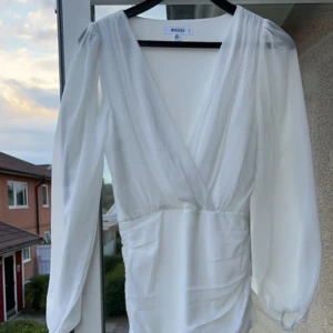 Vit klänning från  Bubbleroom - Elegant vit klänning från Bubbleroom med djup v-ringning och långa, transparenta ärmar. Perfekt för en stilren look.