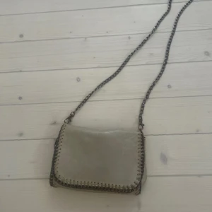 stella mccartney väska - Snygg beige axelväska med kedjedetaljer längs kanterna och en lång kedjerem. Väskan har en praktisk dragkedja inuti och stängs med en tryckknapp. Perfekt för att ge en stilren touch till din outfit.