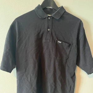 Svart pikétröja från Yoke - Säljer en stilren svart pikétröja från Yoke. Tröjan har korta ärmar och en klassisk krage med knappar. På bröstet finns en praktisk ficka med Yoke-logga. Perfekt för en avslappnad look.