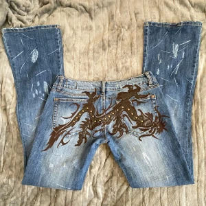 Blå jeans med broderad design - Unika blå jeans med en cool broderad drak design på bakfickorna. Jeansen har en sliten look med ljusa slitningar och en bootcut-stil. Jeansen är lågmidjade och bootcut. ❌Har INGA bilder på❌ mått : midjemått: ca 86 innerbenslängd: ca 82