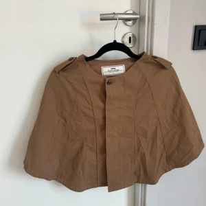 Beige cape från Monki - Snygg beige cape från Monki med knappdetaljer. Perfekt för en stilren look. Den har en enkel design med en knapp framtill och axelremmar som ger en unik touch.