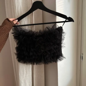 Svart volang tubtopp - Endast använd en gång. Säljer en svart tubtopp med volangdetaljer. Toppen är axelbandslös och har en fluffig design som ger en festlig look. Perfekt för att sticka ut på festen!