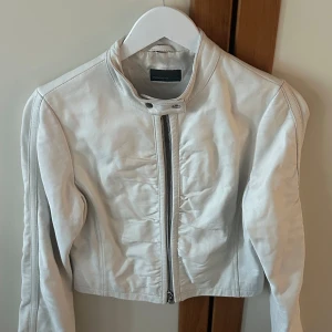 Vit skinnjacka från Rock'n Blue - Snygg vit skinnjacka från Rock'n Blue med dragkedja framtill och en stilren design. Jackan har en hög krage med tryckknappar och långa ärmar. Perfekt för att ge din outfit en cool touch.