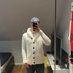Vit stickad kofta med knappar - Säljer en snygg vit stickad kofta från Ralph Lauren. Den har en mysig huva och stora bruna knappar framtill. Fodrad. Storlek M men sitter lite kort på mig som är 183cm. Ny pris ligger lite över 3000kr