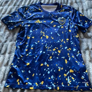 Blå fotbollströja från Adidas - Säljer en blå fotbollströja från Adidas med ett färgglatt konfettimönster i gult och ljusblått. Tröjan har korta ärmar och ett klubbmärke på bröstet. Perfekt för fotbollsfans som vill sticka ut på läktaren eller planen.