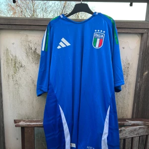 Blå fotbollströja från Adidas - Säljer en blå fotbollströja från Adidas med Italiens landslagsemblem på bröstet. Tröjan har korta ärmar och vita detaljer på sidorna. Perfekt för fotbollsfans som vill visa sitt stöd för Italien.