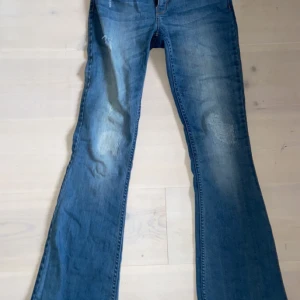 Blå bootcut jeans - Snygga blå bootcut jeans med slitna detaljer på framsidan och bakfickor.Klassisk femficksdesign och knappgylf.