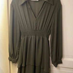  klänning från Zara - Elegant olivgrön klänning med v-ringning och guldknappar framtill. Den har långa ärmar och ett smockat midjeparti som ger en fin passform. Från Zara och är använd 2 ggr inga defekter 
