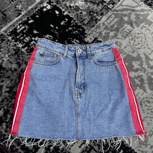 Jeanskjol med röda detaljer från Authentic Denim - Snygg jeanskjol från Authentic Denim med röda och vita ränder på sidorna. Kjolen har en fransig nederkant och klassiska femficksdetaljer. Perfekt för en avslappnad stil.