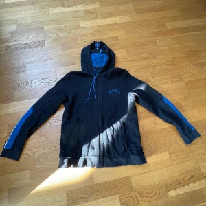 Svart hoodie från Boss - Säljer en svart hoodie från Boss med blå detaljer och dragkedja. Den har en huva med blått foder och snörning. Ärmarna har blå inslag och det finns ett Boss-märke på bröstet. Perfekt för en casual look. Lite skador på armbågen 