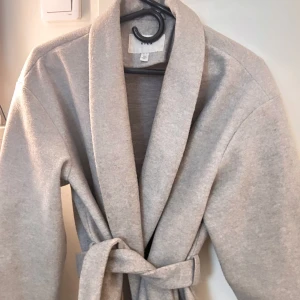 Grå kappa med knytband - Säljer en stilren grå kappa med knytband i midjan. Jackan har en klassisk design med långa ärmar och en mjuk, bekväm passform. Perfekt för att hålla sig varm och snygg under kyligare dagar. Kappan är från ”vila” här inte använt den så många gånger eftersom den står i storleken tycker jag personligen. Annars inga skador så perfekt till våren. 