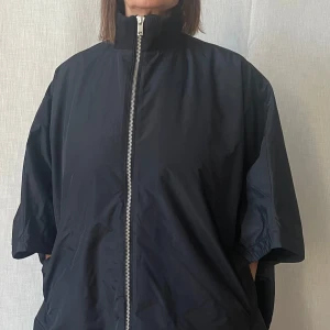 Svart vindjacka med dragkedja - Snygg svart vindjacka med dragkedja och kortärmad design. Jackan har en hög krage och praktiska fickor på sidorna. Storlek one size! Mycket bra skick!