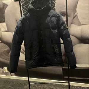 Mörkblå dunjacka från Stone Island Junior - Säljer en snygg mörkblå dunjacka från Stone Island Junior. Jackan har en klassisk design med dragkedja och en avtagbar huva. Den ikoniska loggan sitter på ärmen. Perfekt för kyliga dagar! Den håller värme väldigt bra, den kan användas till höst och vinter jacka men även när man åker skidor. Ny pris 6000kr. Den är i väldigt bra skick fortfarande.