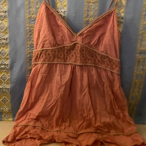 Rosa linne/kort klänning med spetsdetaljer - Charmigt rosa linne/kort klänning med smala axelband och v-ringning. Toppen har vackra spetsdetaljer runt midjan och längs kanterna, vilket ger en romantisk touch. Perfekt för varma dagar.