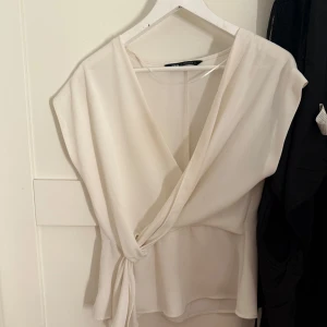 Vit omlottblus från Zara - Elegant vit omlottblus från Zara med knytning i midjan. Blusen har en v-ringad design och är ärmlös. Knappt använd. 