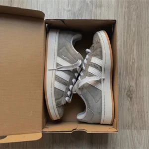 Grå Adidas Campus sneakers - Snygga grå Adidas Campus sneakers i mocka med vita detaljer och klassiska tre ränder på sidan. Skorna har en vit sula och snörning framtill. Perfekta för en stilren och avslappnad look.