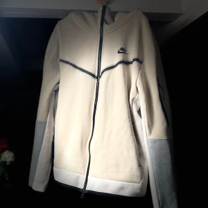 Beige och grå Nike Tech tröja - Säljer en snygg beige huvtröja från Nike med gråa ärmar och dragkedja. Tröjan har en stilren design med Nike-logga på bröstet och en bekväm huva. Perfekt för en avslappnad stil. Funkar som storlek M oxå.