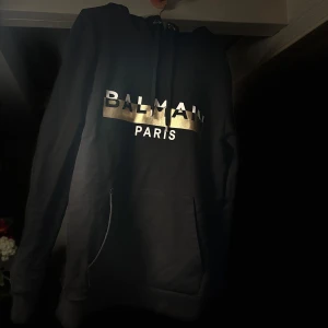 Svart hoodie från Balmain - Snygg svart hoodie från Balmain med guldtryck på bröstet. Den har en klassisk design med en stor ficka framtill och en huva. Perfekt för en stilren och bekväm look.