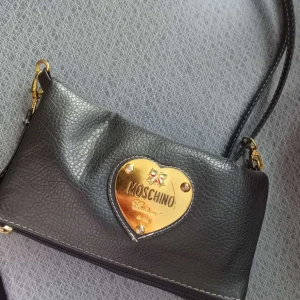 Svart axelväska från Moschino - Snygg svart axelväska från Moschino med ett guldhjärta och logotyp på framsidan. Väskan har en justerbar axelrem och stängs med dragkedja. Perfekt för att ge en touch av lyx till din outfit.