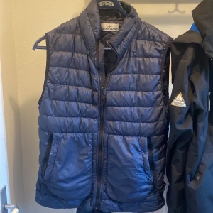 Blå dunväst från Stone Island - Säljer en snygg blå dunväst från Stone Island. Västen har en quiltad design och dragkedja framtill. Perfekt för att hålla värmen under kyliga dagar. Märkeslogga på sidan ger en extra touch av stil.