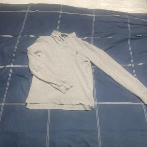 Grå pikétröja från Ralph Lauren - Säljer en stilren grå pikétröja från Ralph Lauren i slim fit. Tröjan har långa ärmar och klassisk krage med knappar. Den ikoniska loggan är broderad på bröstet. Perfekt för en avslappnad men ändå elegant look.