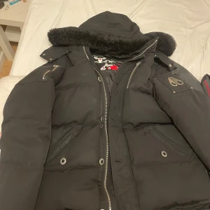 Svart dunjacka från Moose Knuckles - Säljer en svart dunjacka från Moose Knuckles med pälskantad huva och dragkedja. Jackan har flera fickor med dragkedjor och knappar för extra stil och funktionalitet. Perfekt för kalla dagar, är i nyskick och knappt använd. Pris går att diskutera 
