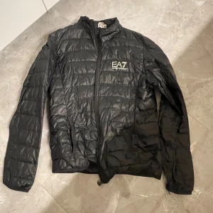 Svart pufferjacka från Emporio Armani - Snygg svart pufferjacka från Emporio Armani EA7. Jackan har en dragkedja framtill och är långärmad. Perfekt för kyligare dagar med sin stilrena design och lätta material. Storlek L men sitter som M, inga defekter förutom att den för tillfället är lite skrynklig.