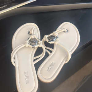 Vita sandaler från Michael Kors - Snygga vita sandaler från Michael Kors med dekorativa metallringar och logotypdetaljer. Sandalerna har en stilren design med flätade remmar och en bekväm sula. Perfekta för varma sommardagar.